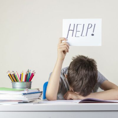 Sad tired frustrated boy sitting at the table with many books and holding help sign. Learning difficulties, education concept.